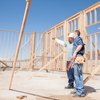Two men in construction gear inspecting the wooden framing of a building on a construction site under a clear blue sky.