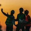 Silhouettes of three construction workers wearing safety helmets and vests, discussing project plans against an orange sunset background.