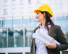 A smiling woman wearing a yellow construction helmet holding rolled-up blueprints outside a modern office building.