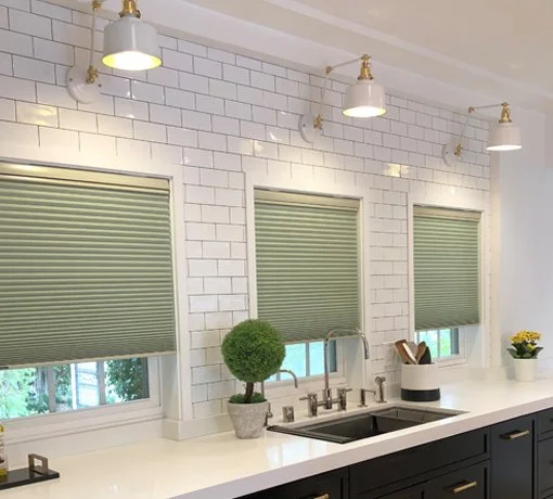 Kitchen with three windows, white subway tile backsplash, black lower cabinets, and white countertop. Green roller blinds cover the windows. Decor includes potted plants, a dish drying rack, and ceiling-mounted light fixtures.