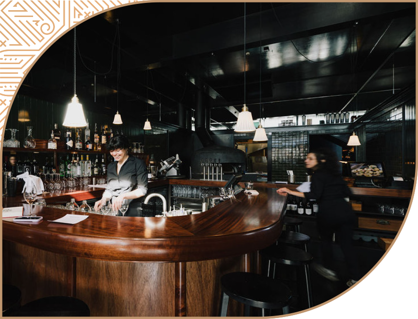 A bartender preparing drinks at a curved wooden bar in a dark, modern restaurant or bar setting with hanging pendant lights.
