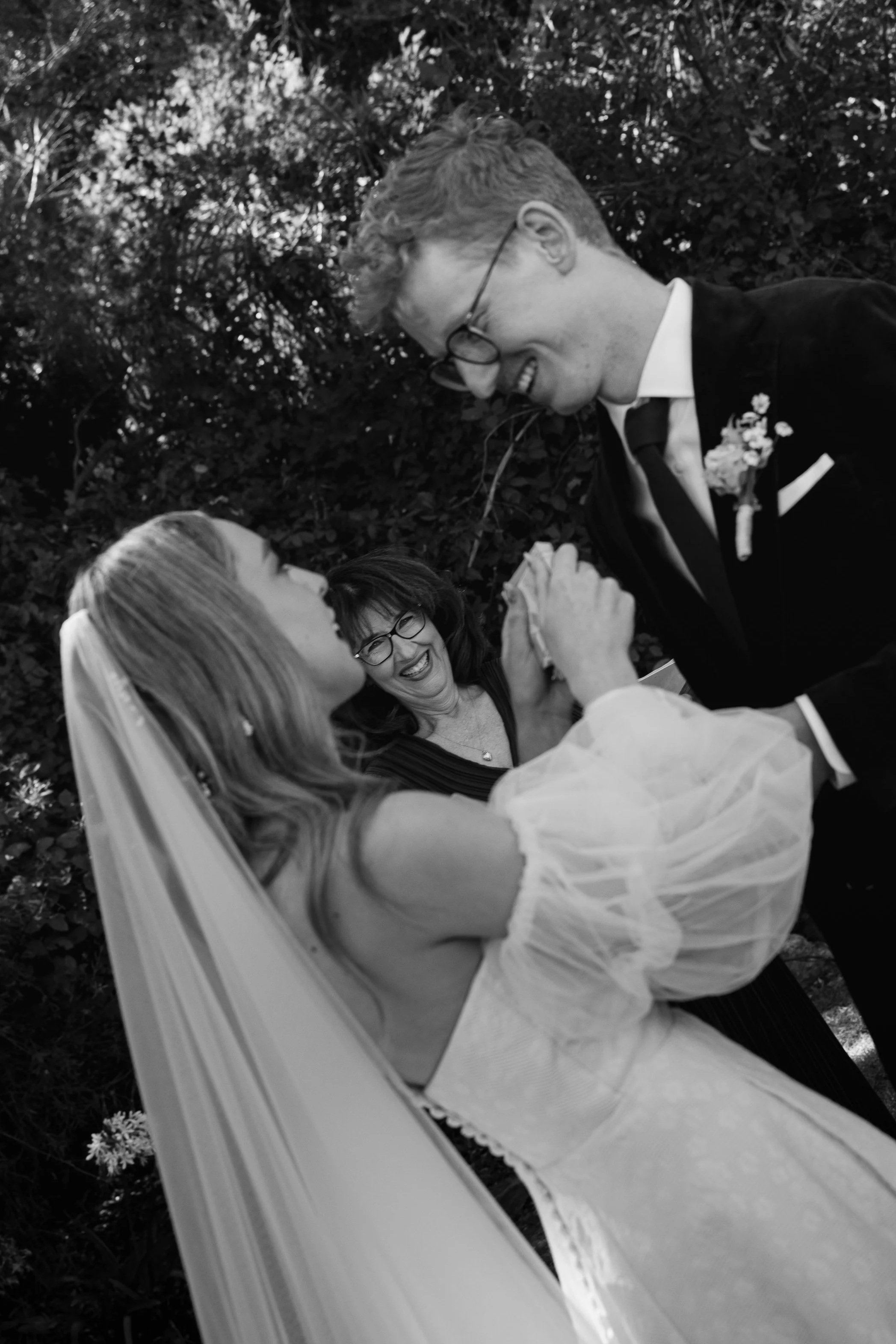 A wedding ceremony outdoors with a bride and groom holding hands and looking at each other, while a woman in glasses smiles in the background.