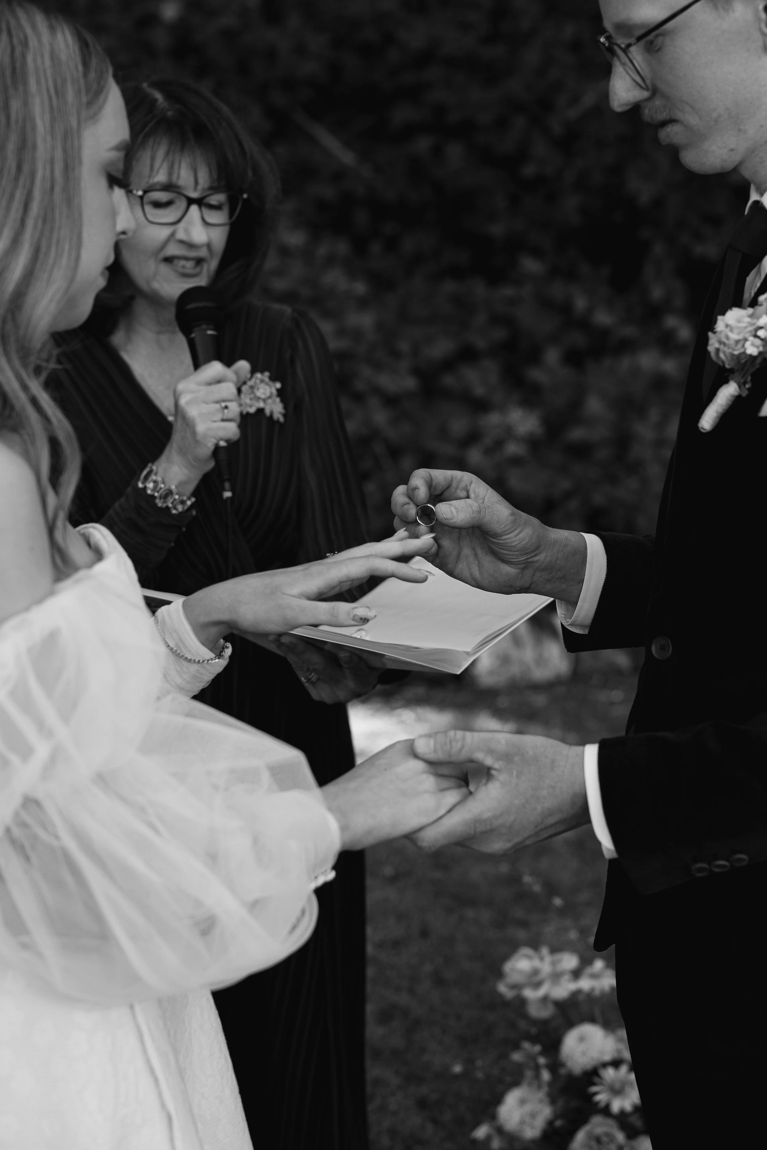 A black-and-white photo of a wedding ceremony where a bride and groom exchange rings. The officiant, holding a microphone, reads from a paper as she presides over the ceremony outdoors with flowers in the background.