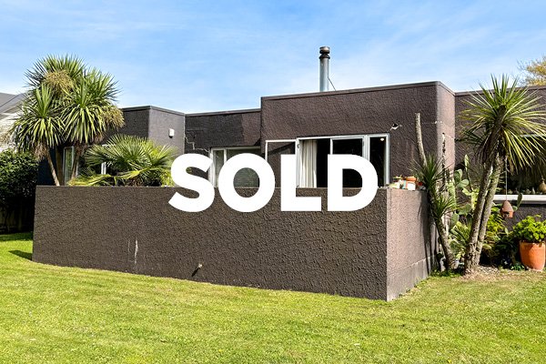 Modern brown house with a large «SOLD» sign on the front yard, tropical plants, and a well-maintained green lawn.
