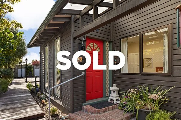 Exterior of a house with a dark wooden facade, red front door, large front window, and some greenery, with a white sold sign in the foreground.