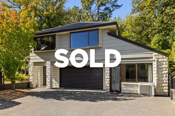 A modern two-story house with a stone exterior, black garage door, large front window, and surrounded by green trees, with the word SOLD over the image.