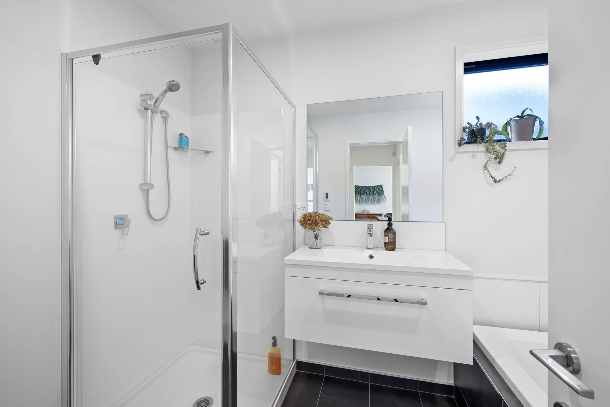 Modern bathroom with a glass shower enclosure, a white vanity with a mirror, a small window with potted plants, and a black tiled floor.