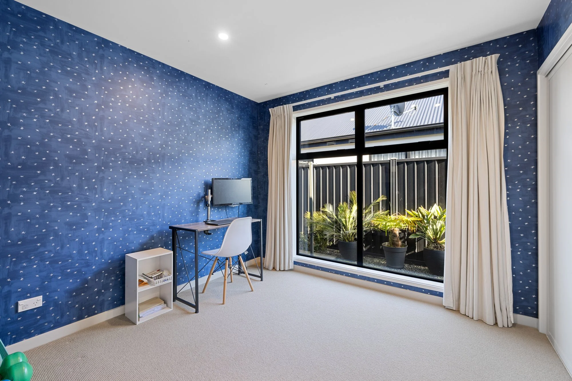 A bedroom with blue star-patterned wallpaper, a large window with white curtains, a small desk with a computer, a white chair, a small white bookshelf, and potted plants outside the window.
