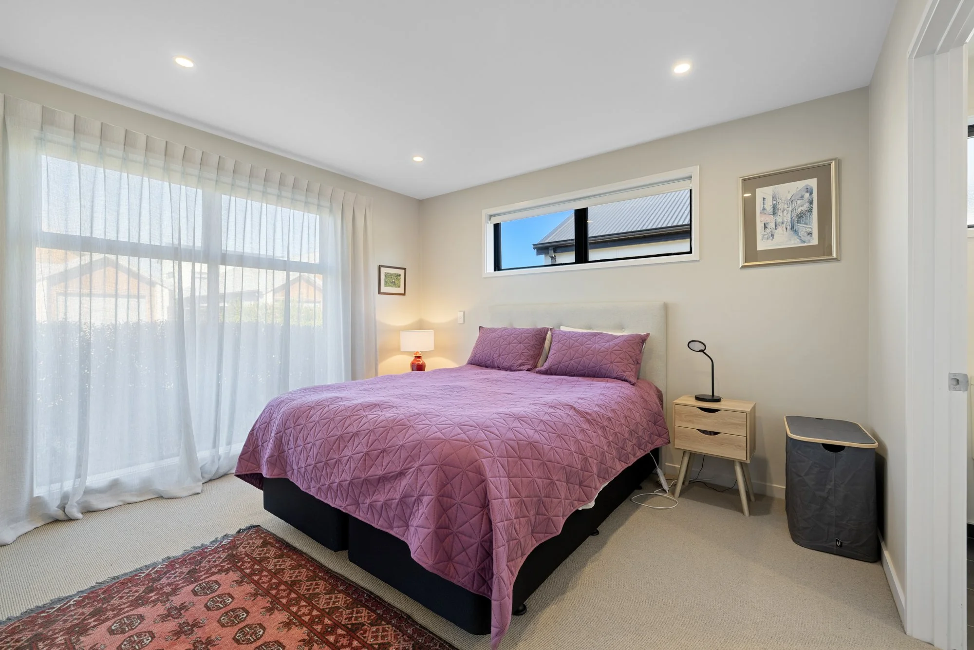 A bedroom with a bed covered in a pink quilt and pillows, a nightstand with a lamp, a window above the bed, and curtains on a large window to the left.