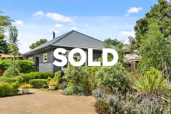 A house with a gray roof partially hidden behind lush green shrubs and plants, with a path leading up to it, under a bright blue sky with some clouds, and large white text saying "SOLD" across the image.