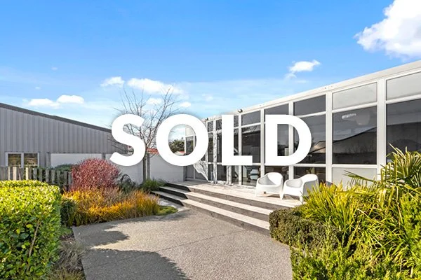 Modern building with outdoor seating, bushes, and a clear blue sky with clouds.