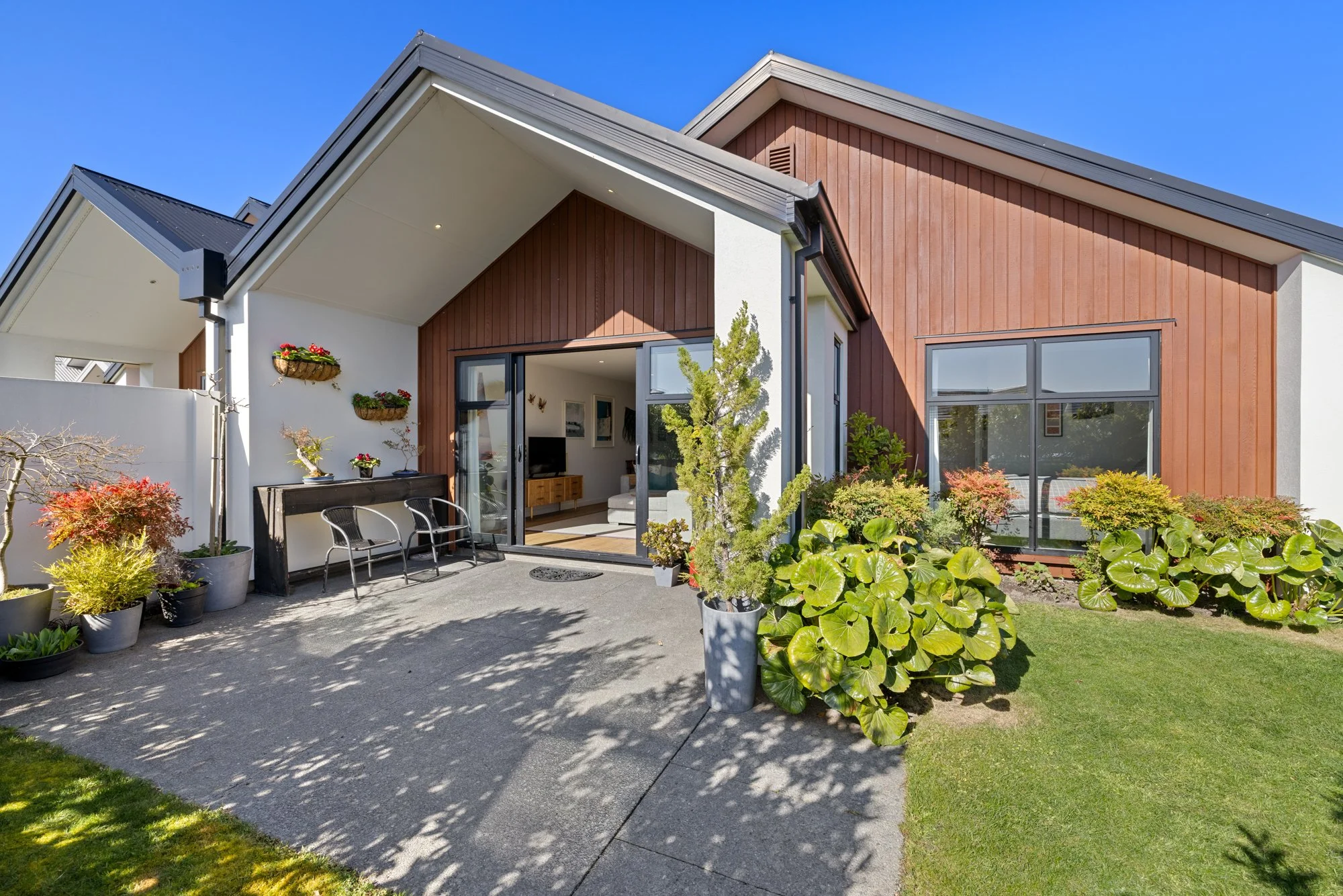 Modern house with an outdoor patio, sliding glass doors, and a garden with green plants and colorful shrubs under a sunny blue sky.