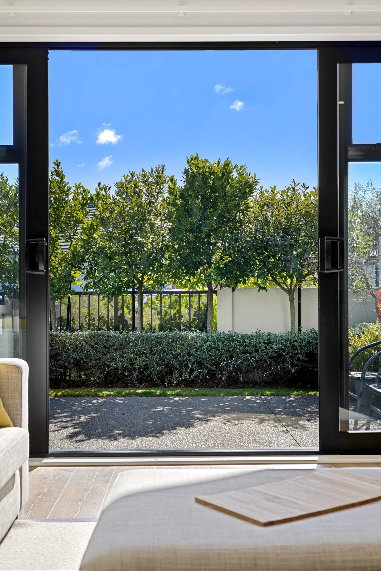 Interior view from inside a house looking out through open sliding glass doors at a sunny backyard with trees, bushes, a white fence, and a patio surface, under a partly cloudy blue sky.
