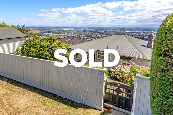 View of a residential backyard with a white wooden fence, gate, green shrubbery, of sold house with rooftops and cityscape in the background under cloudy sky.