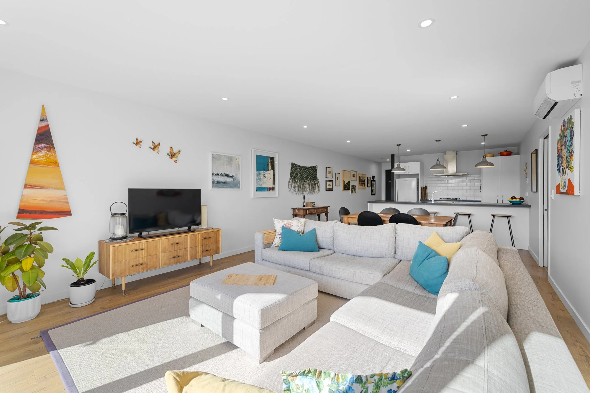 Bright living room with white walls, a beige sectional sofa with colorful cushions, a wooden TV stand with a flat-screen TV, potted plants, and artwork on the wall; open kitchen with white cabinets, a breakfast bar with stools, and pendant lighting.