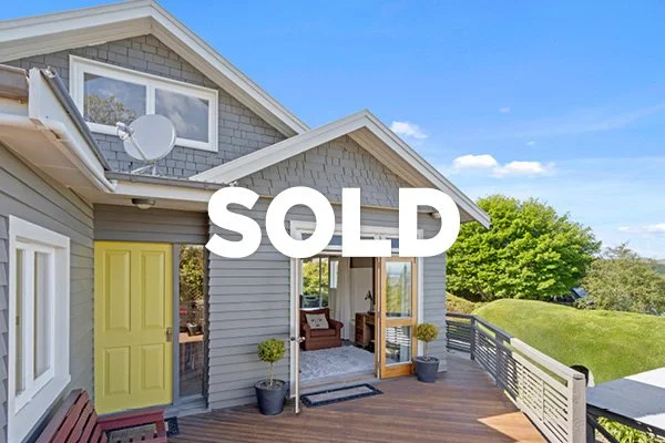 A house with a wooden deck, yellow door, and large sliding glass doors, with a bright blue sky and green trees in the background. The word 'SOLD' is overlaid on the image.