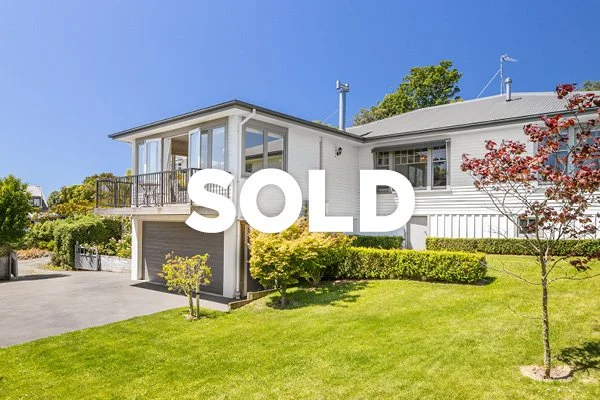 A sold sign hangs in front of a modern white house with a balcony, surrounded by green lawn and trees.