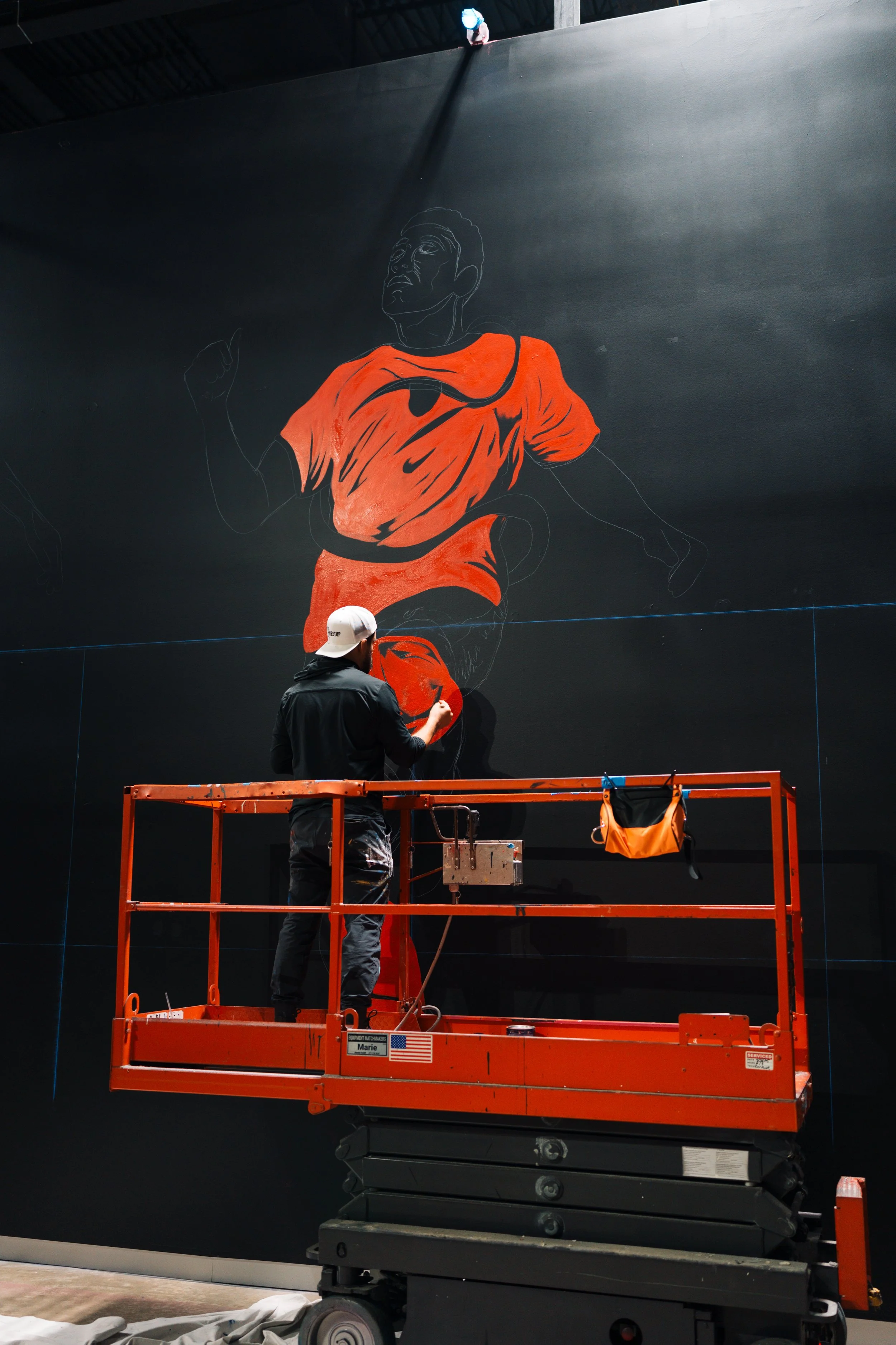 An artist is painting a large mural on a dark wall. The mural depicts a man running, wearing an orange shirt with a Nike logo. The artist is standing on a red lift, working on the lower part of the mural.