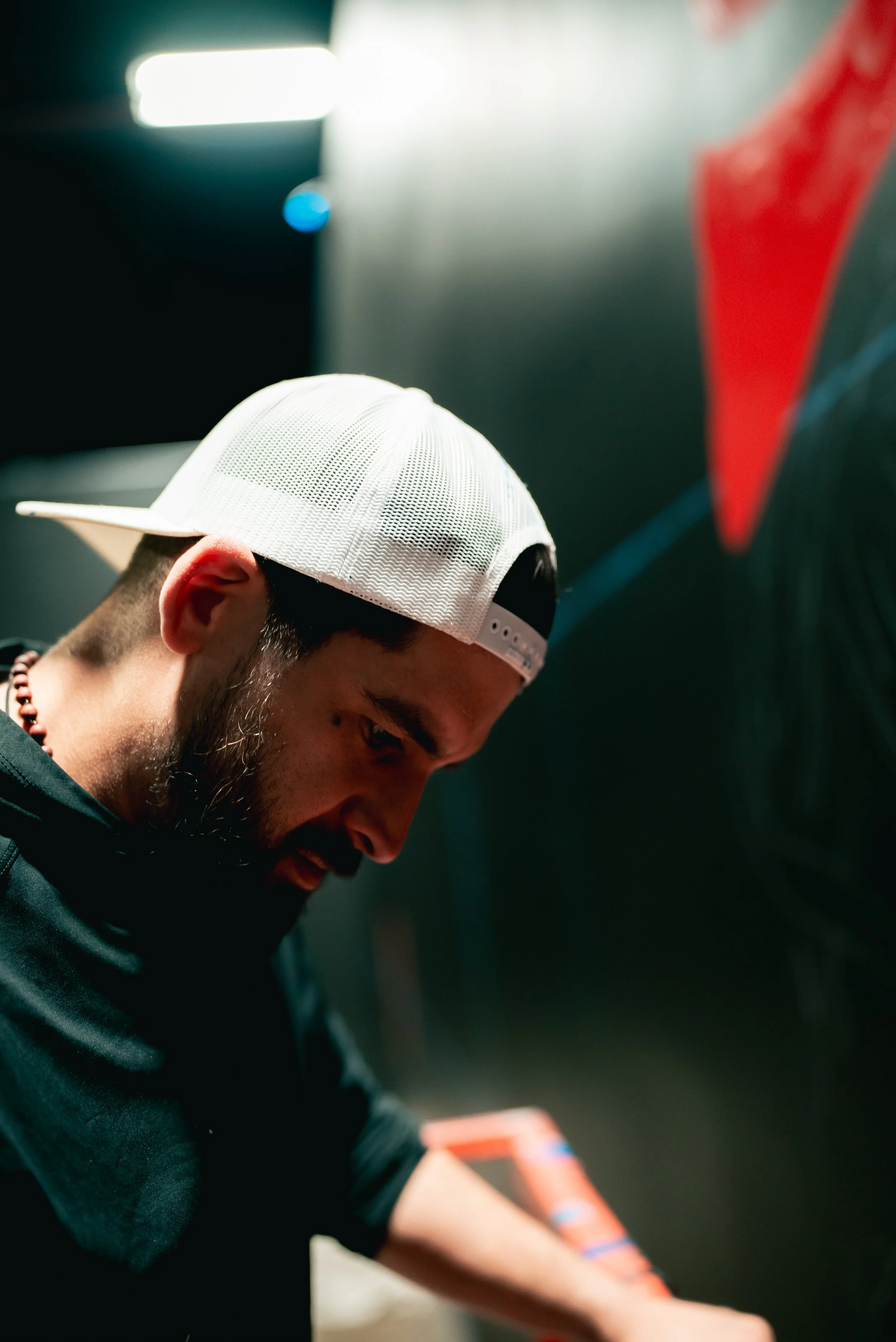 A man wearing a white baseball cap and black shirt, looking down, with a blurred background that includes a red and black object.