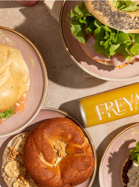 A tray of breakfast foods including a bagel, a sandwich with lettuce and ham, a bowl of scrambled eggs, and a can of orange juice.