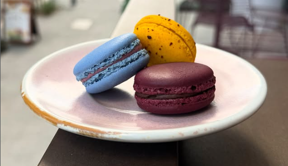 Three colorful macarons on a pink plate, with a yellow, a blue, and a purple macaron, on a dark surface outside.