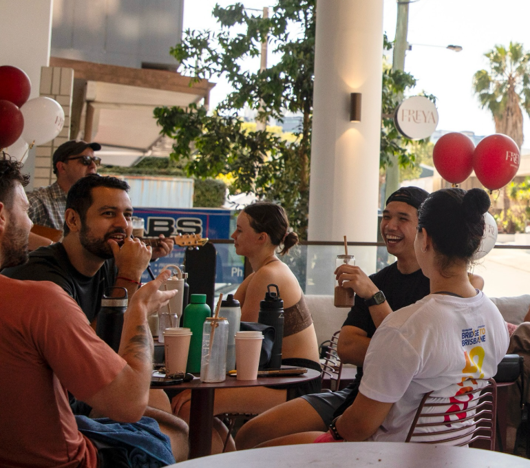 Group of young adults socializing and laughing at an outdoor cafe, with drinks and water bottles on the table, balloons and trees in the background.