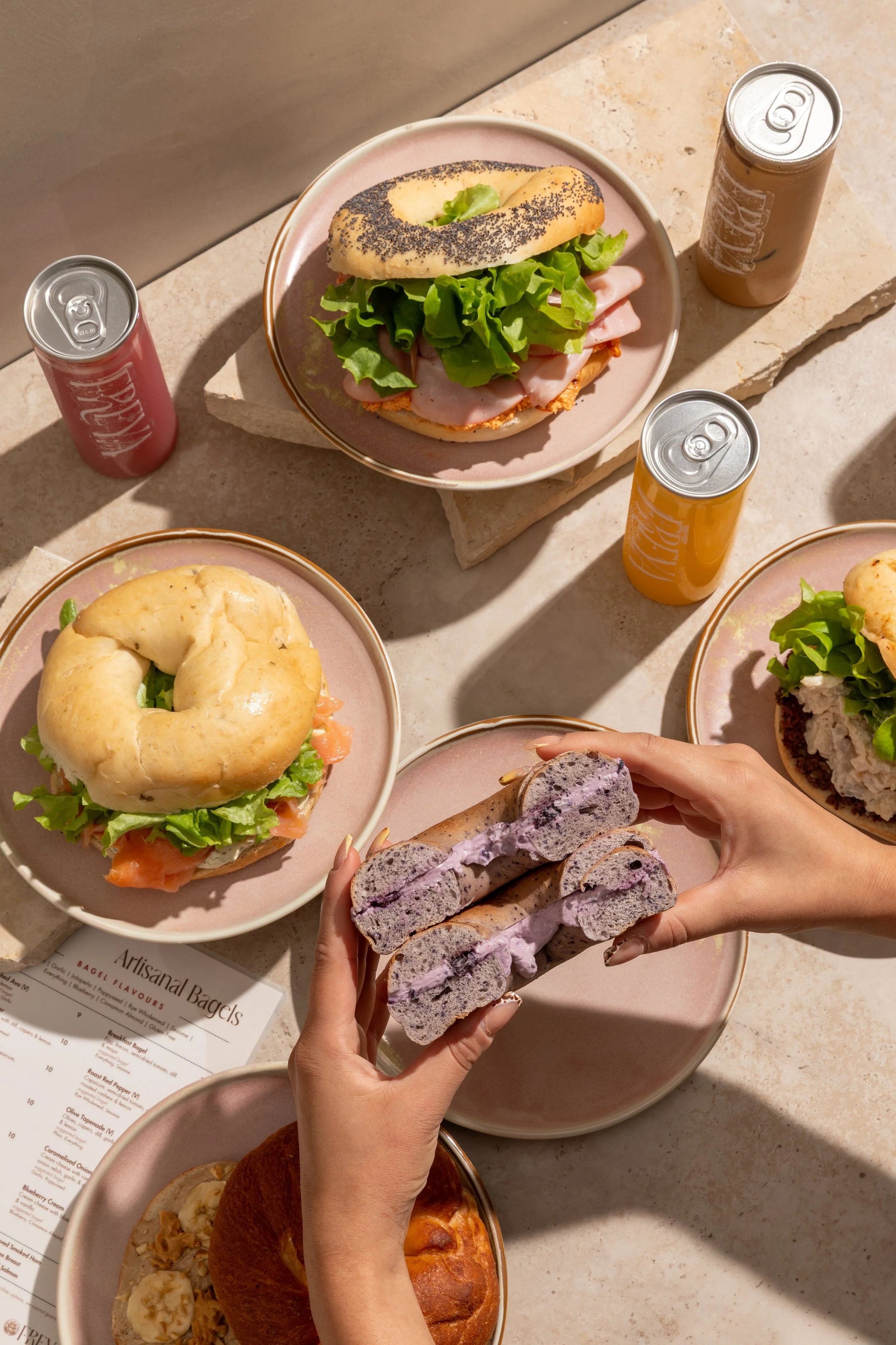 A table with three plates of bagels and sandwiches, three cans of soda, and a menu, with a person's hands holding purple ice cream cookies.