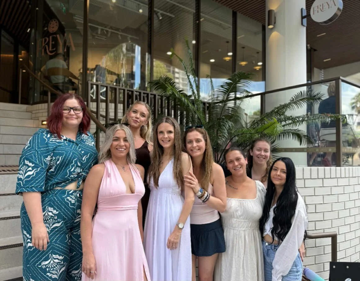 Group of eight women standing in front of a restaurant entrance, smiling. The women are dressed in casual and summer dresses, standing on stairs outside near a large potted plant.