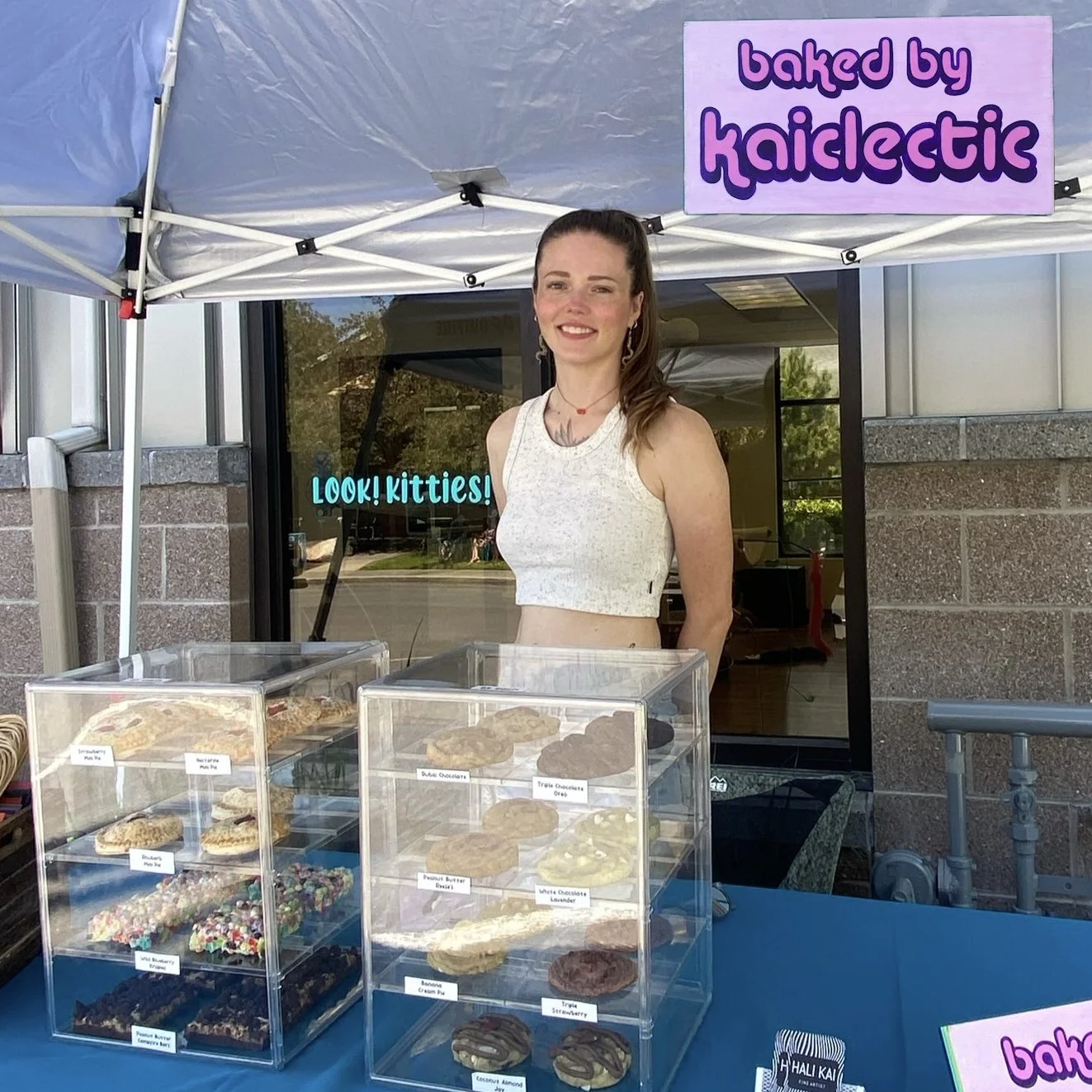 A woman standing behind a display of cookies at a baked goods booth under a tent, with a sign reading 'baked by kaiclectic'.