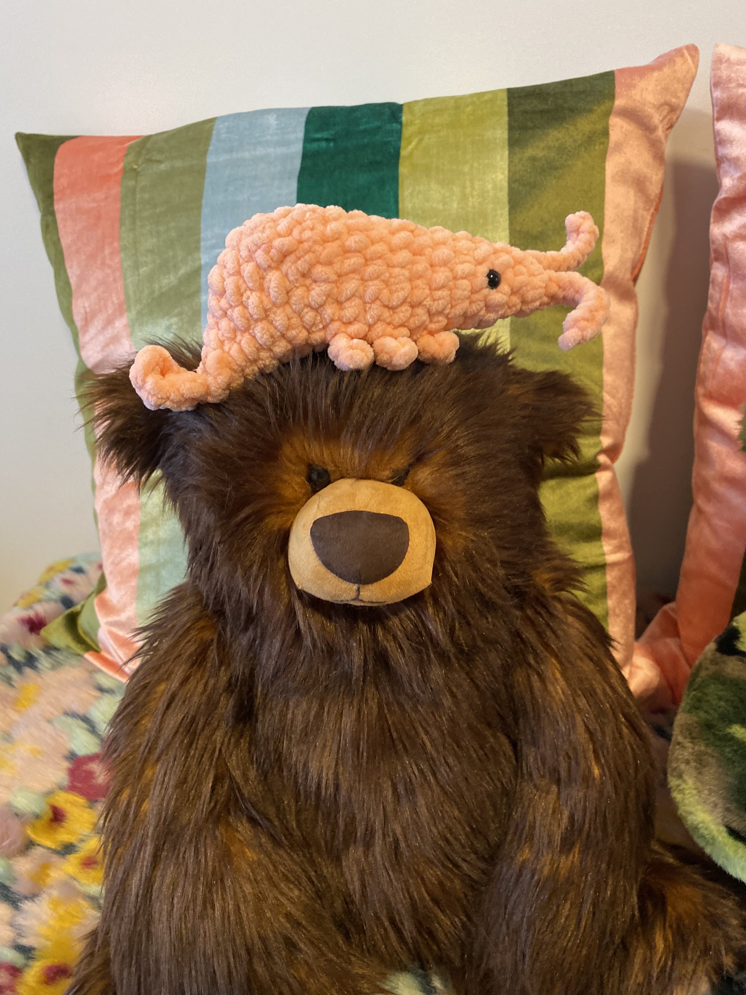 A brown bear stuffed animal with a crocheted pink shrimp plushie on his head.