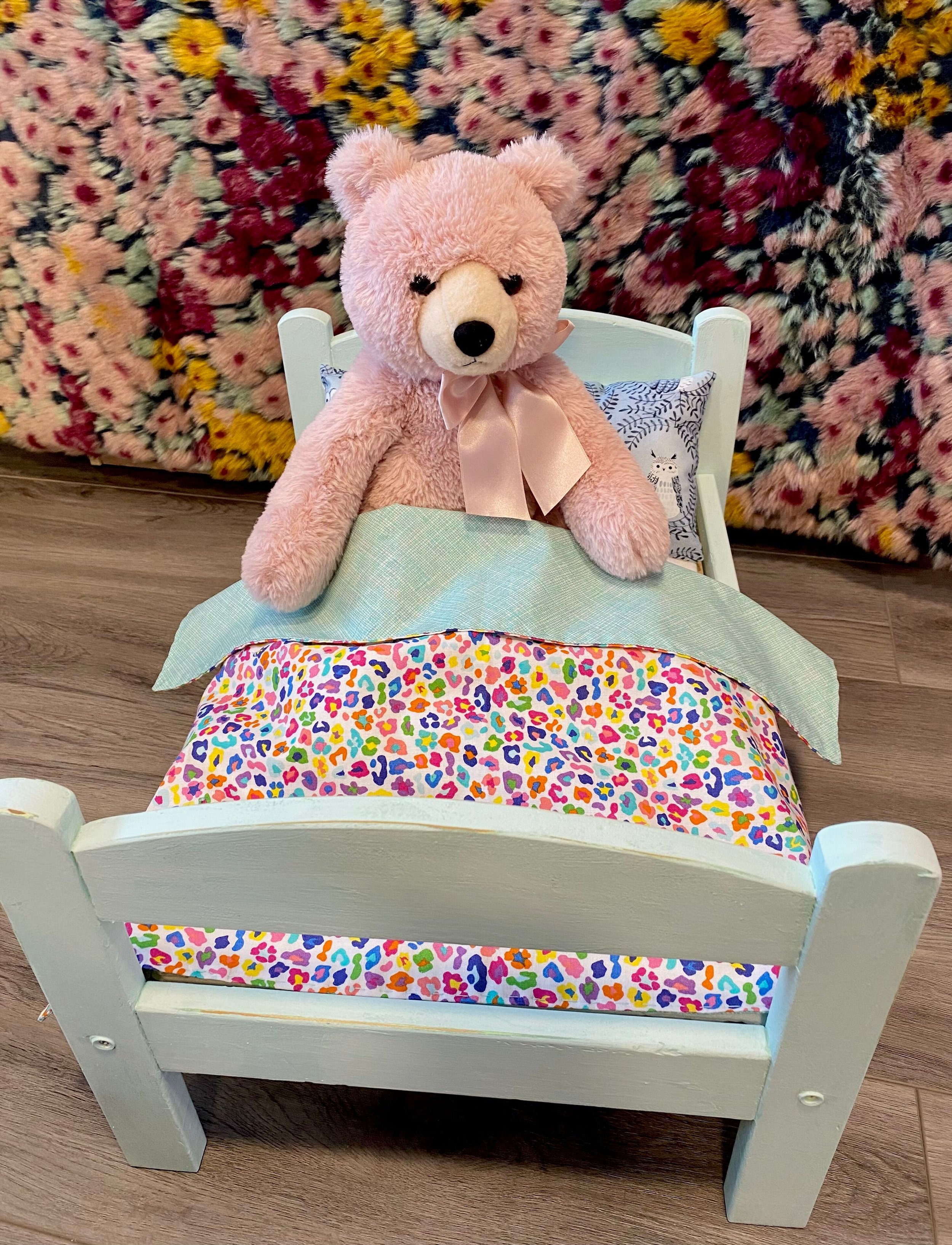A pink teddy bear in a toy bed with a blanket and pillow.