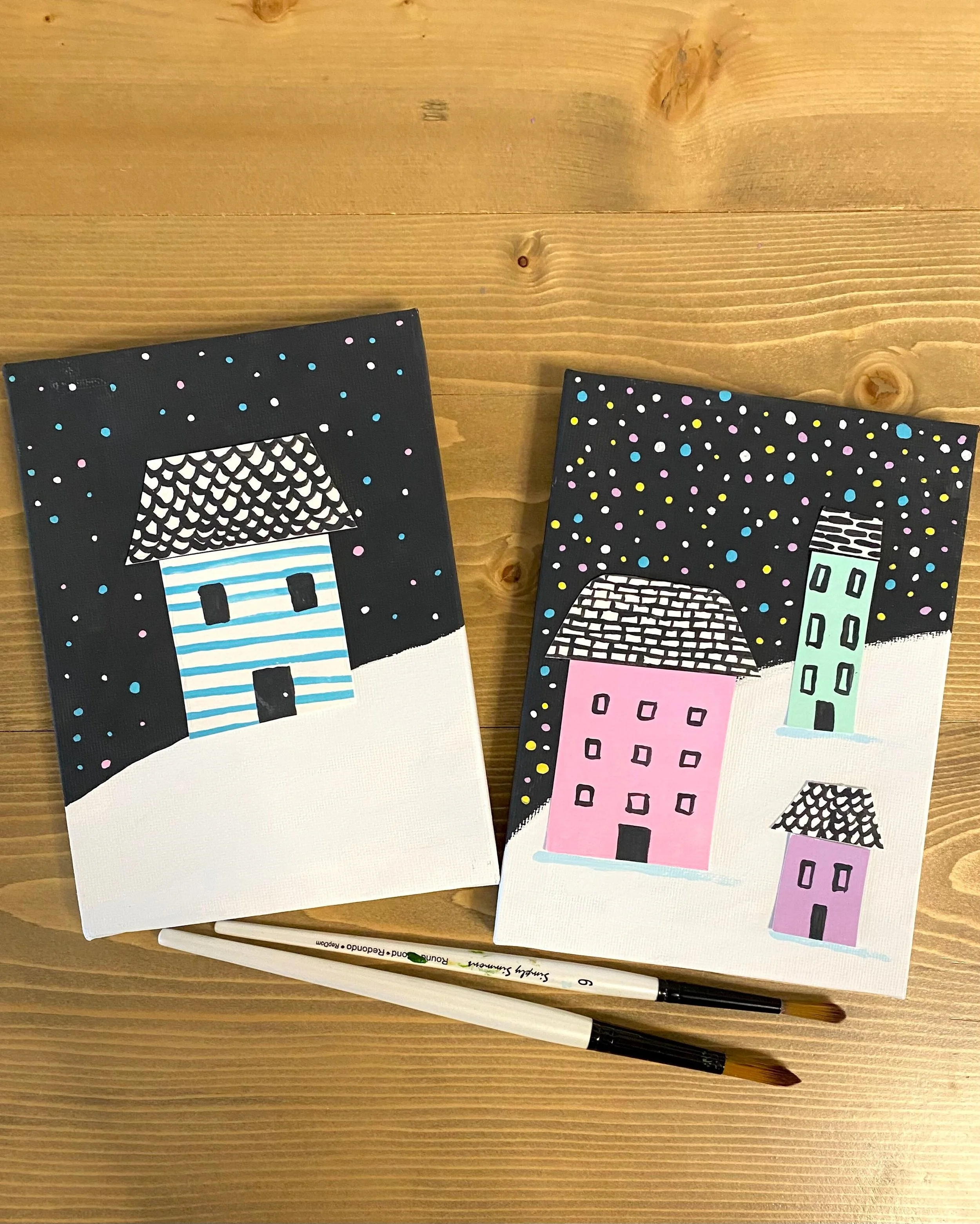 Two small paintings of houses in the winter on a wood table.