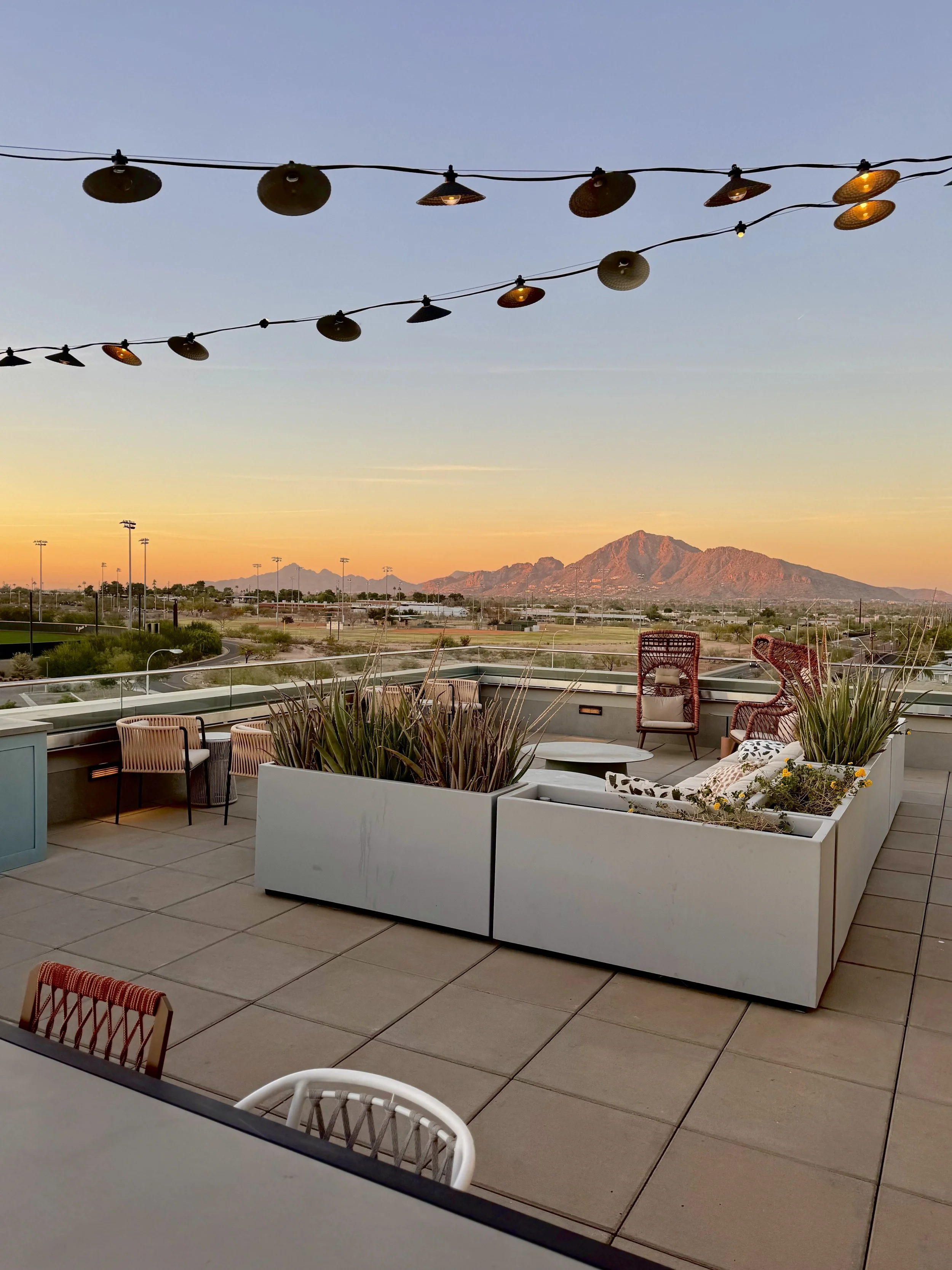 Rooftop terrace with outdoor seating including chairs, a sofa, and large planters, overlooking a desert landscape with mountains at sunset, and string lights hanging overhead.