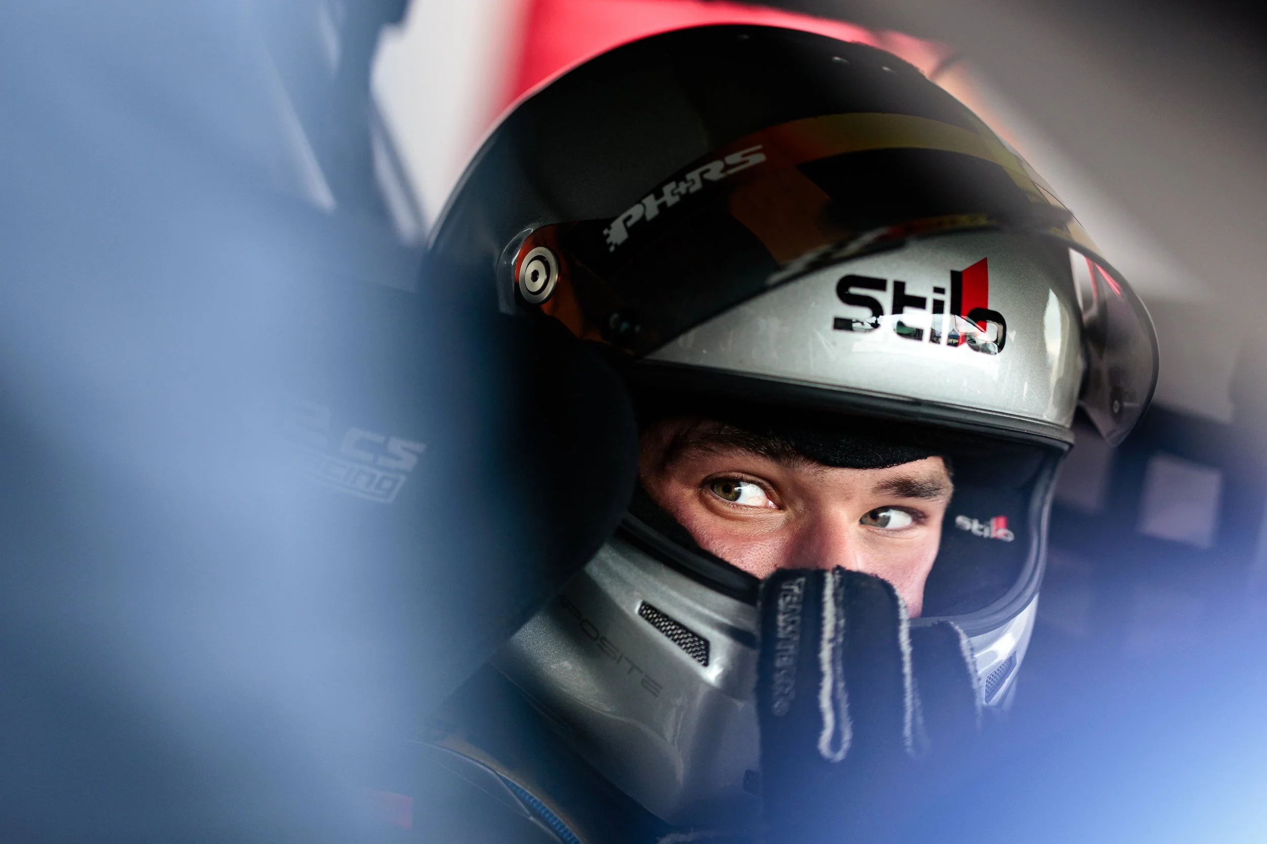 A race car driver wearing a helmet with the brand 'Stilo', sitting in a race car, looking to the side with focused eyes.