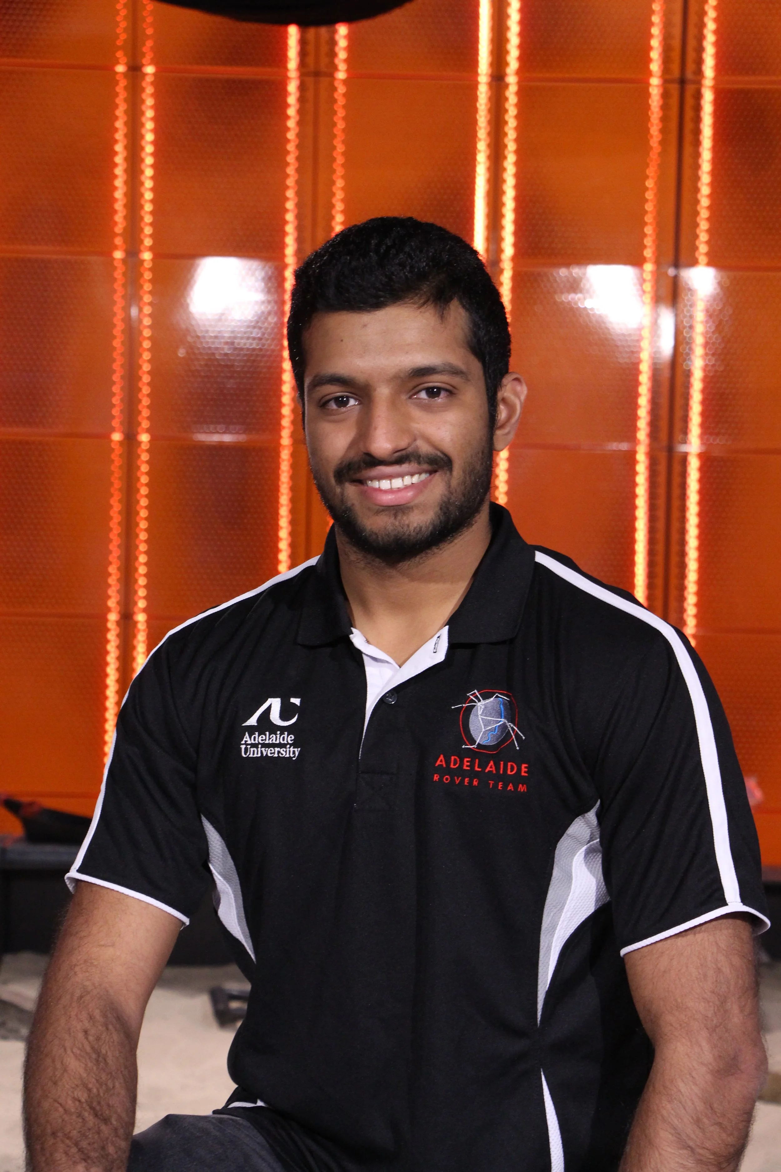 A smiling young man with dark hair in a dark navy blue polo shirt, with an Adelaide Rover Team Logo on his right chest-front and an Adelaide University logo on his left chest-front, smiling indoors against an orange L.E.D. background in a tech lab.