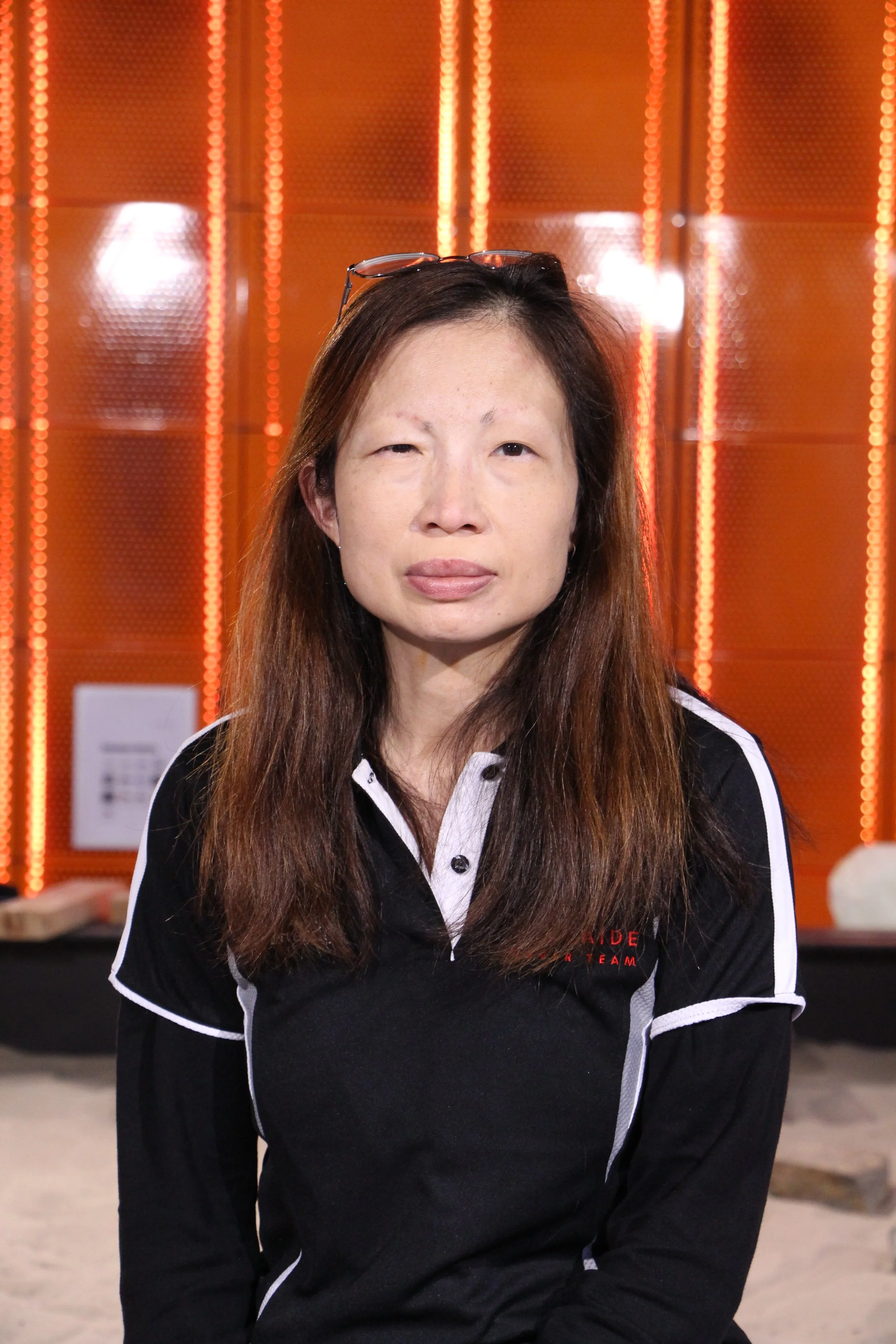 A woman in a dark navy blue polo shirt, with glasses on her forehead and long auburn hair flowing in front what seems to be  an Adelaide Rover Team Logo, standing indoors against an orange L.E.D. background in a tech lab.