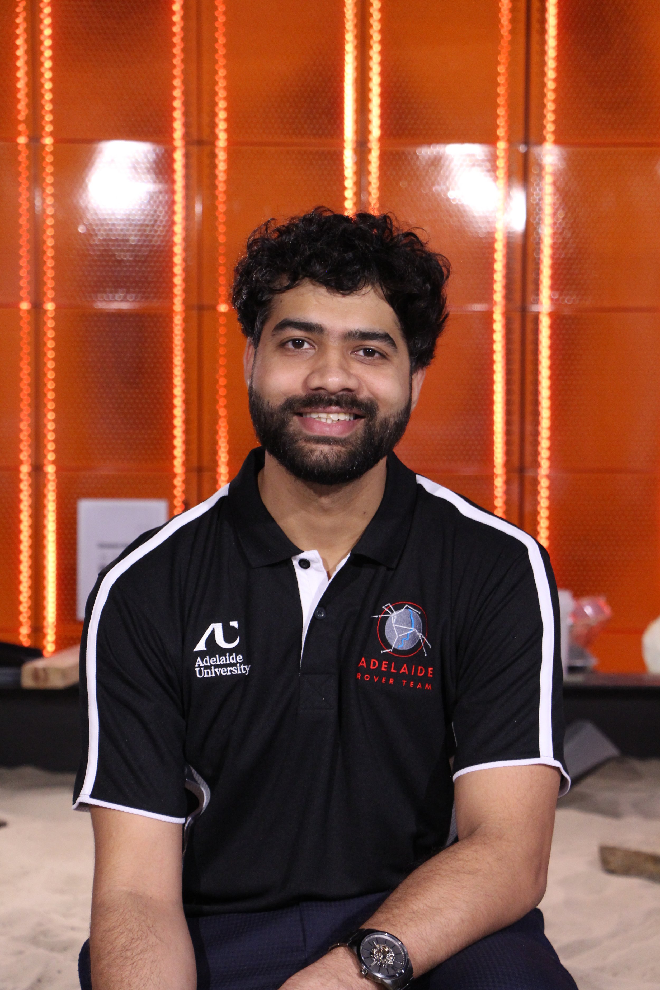 A smiling young man with dark hair in a dark navy blue polo shirt, with an Adelaide Rover Team Logo on his right chest-front and an Adelaide University logo on his left chest-front, standing indoors against an orange L.E.D. background in a tech lab.