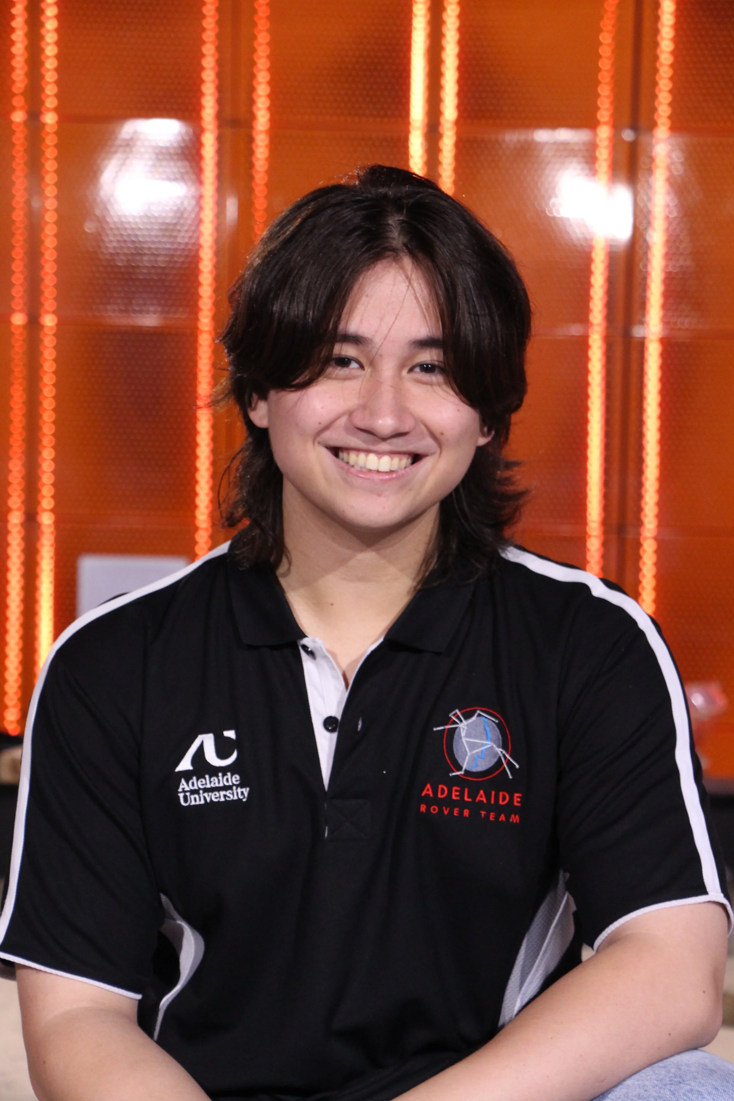 A smiling young man with dark hair in a dark navy blue polo shirt, with an Adelaide Rover Team Logo on his right chest-front and an Adelaide University logo on his left chest-front, smiling indoors against an orange L.E.D. background in a tech lab.