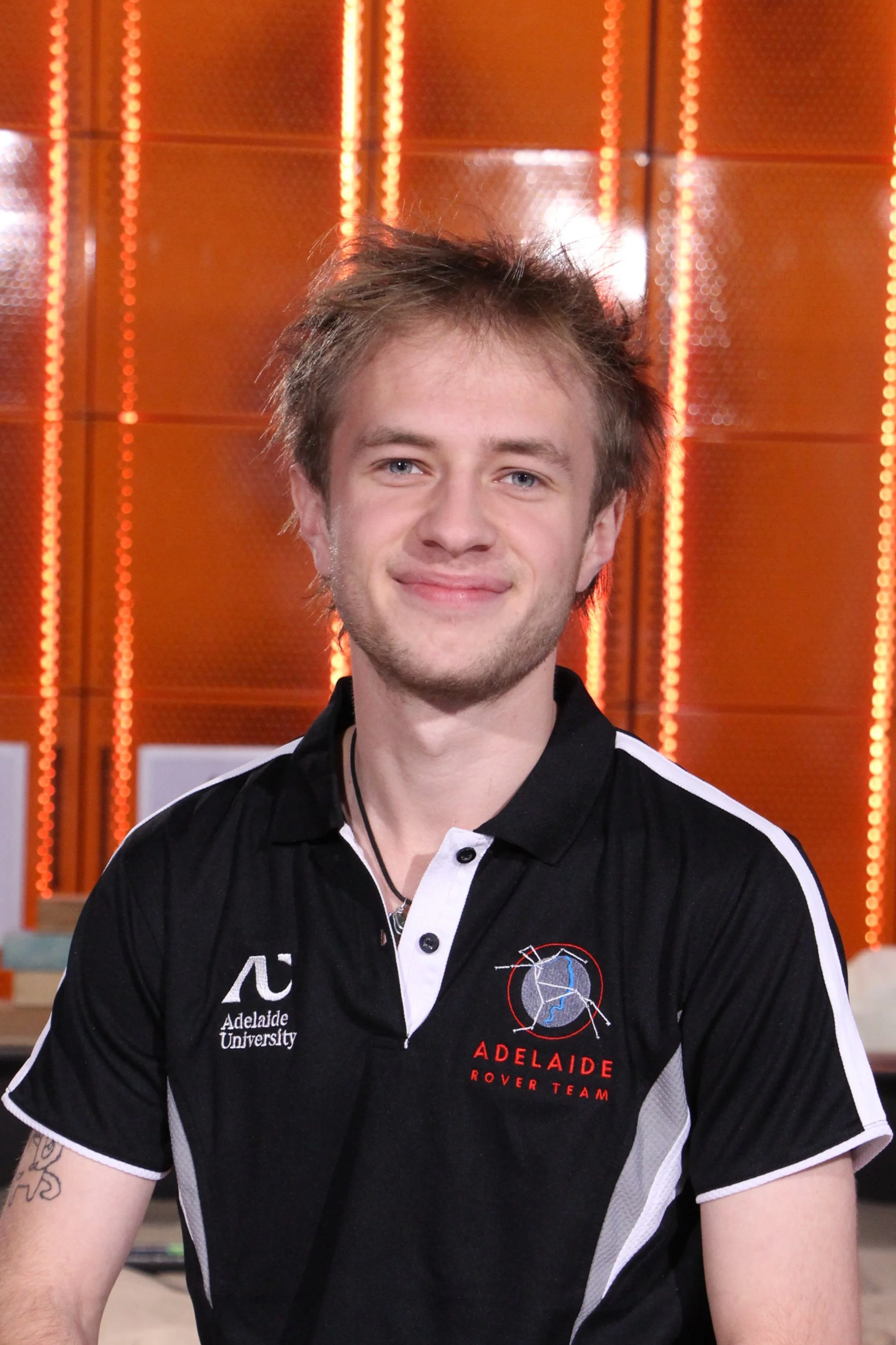 A smiling young man with blonde hair in a dark navy blue polo shirt, with an Adelaide Rover Team Logo on his right chest-front and an Adelaide University logo on his left chest-front, smiling indoors against an orange L.E.D. background in a tech lab.