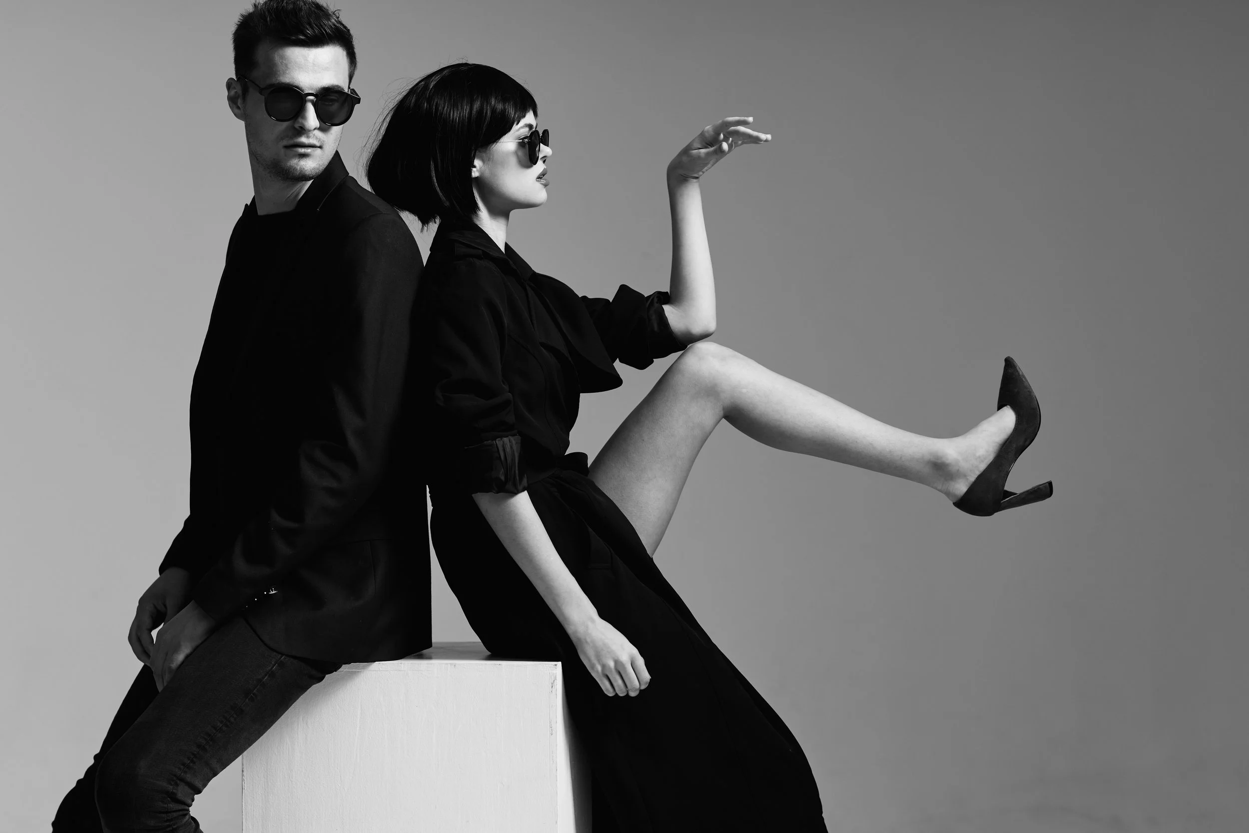 Black and white fashion photo of two models, a man and a woman, posing back-to-back. They are wearing sunglasses and stylish black clothing. The woman has one leg raised, wearing high heels.