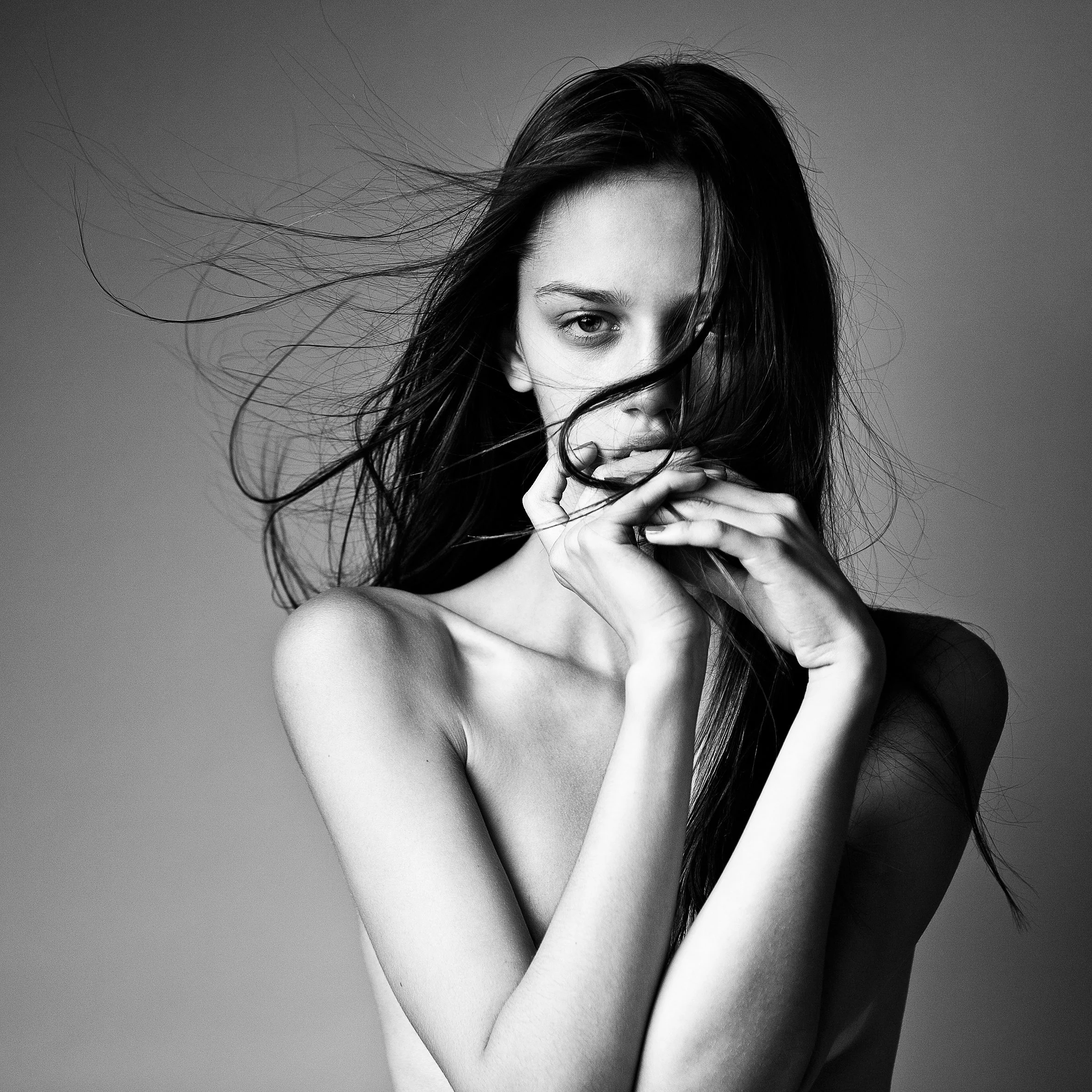 Black and white portrait of a person with long hair covering part of their face, looking at the camera, with their hands near their mouth.