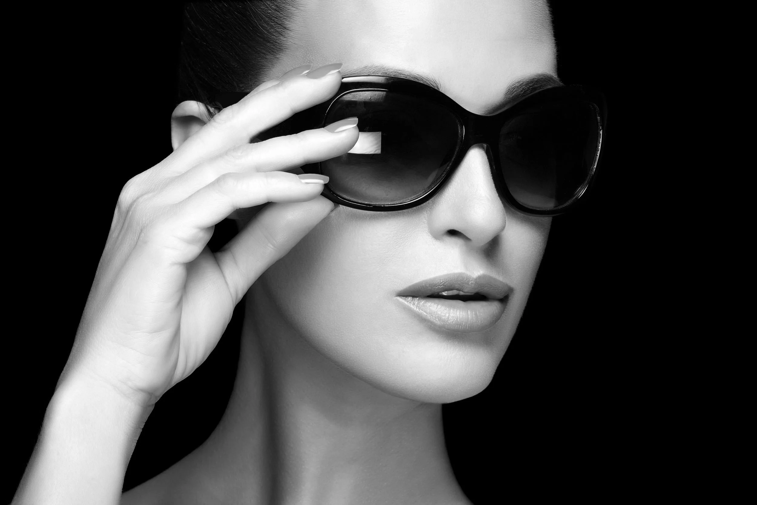 Black and white photo of a woman wearing sunglasses, holding one side with her fingers against a dark background.