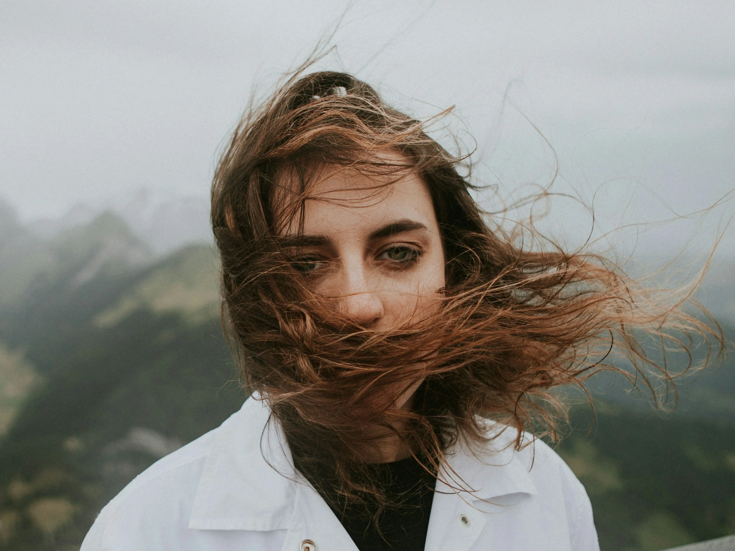 A woman with brown hair blowing in the wind, wearing a white jacket, with a mountain landscape in the background.