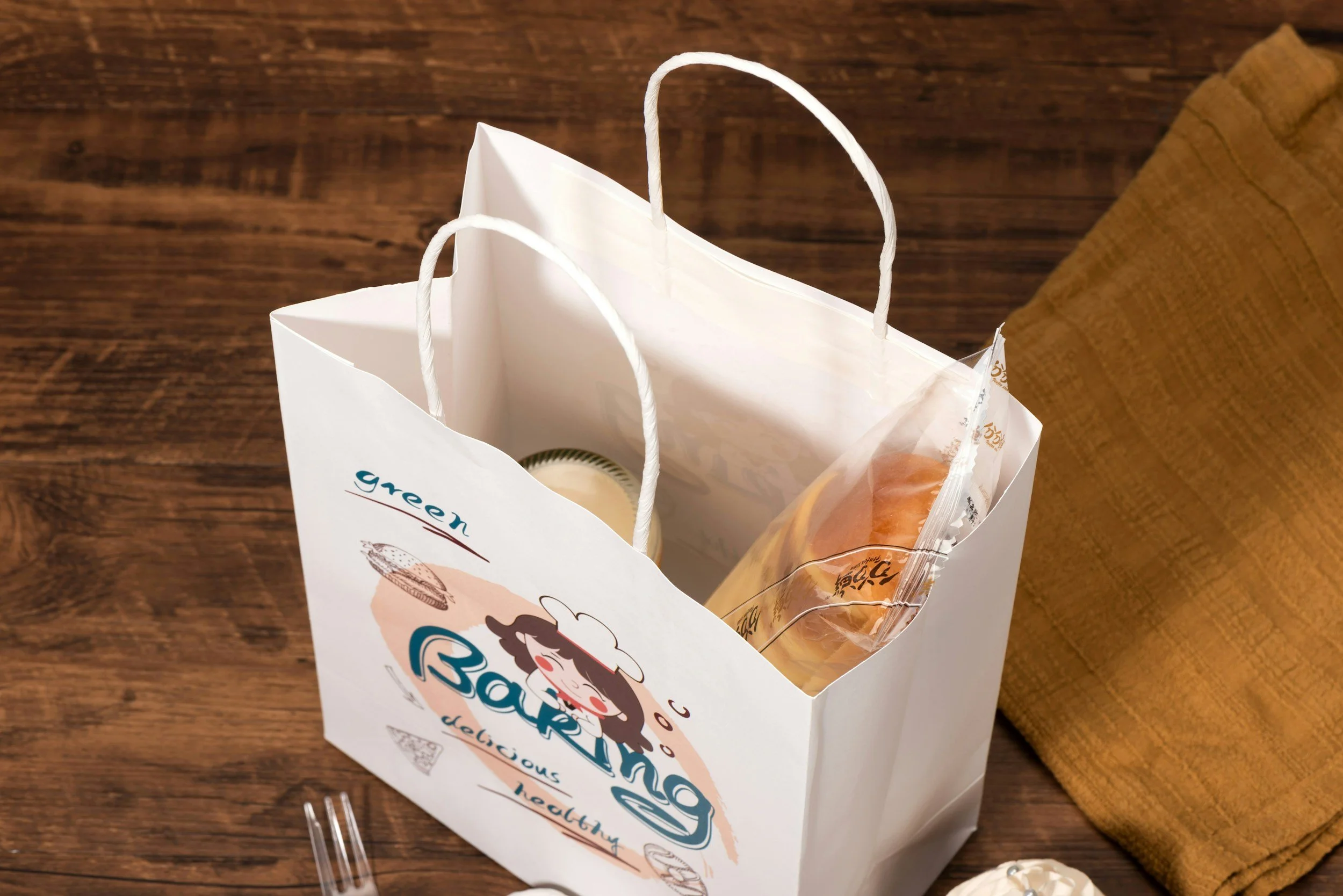 A white paper takeout bag with a cartoon girl chef illustration, containing a plastic cup, a wrapped sandwich, and other items, on a wooden table with a brown cloth napkin nearby.