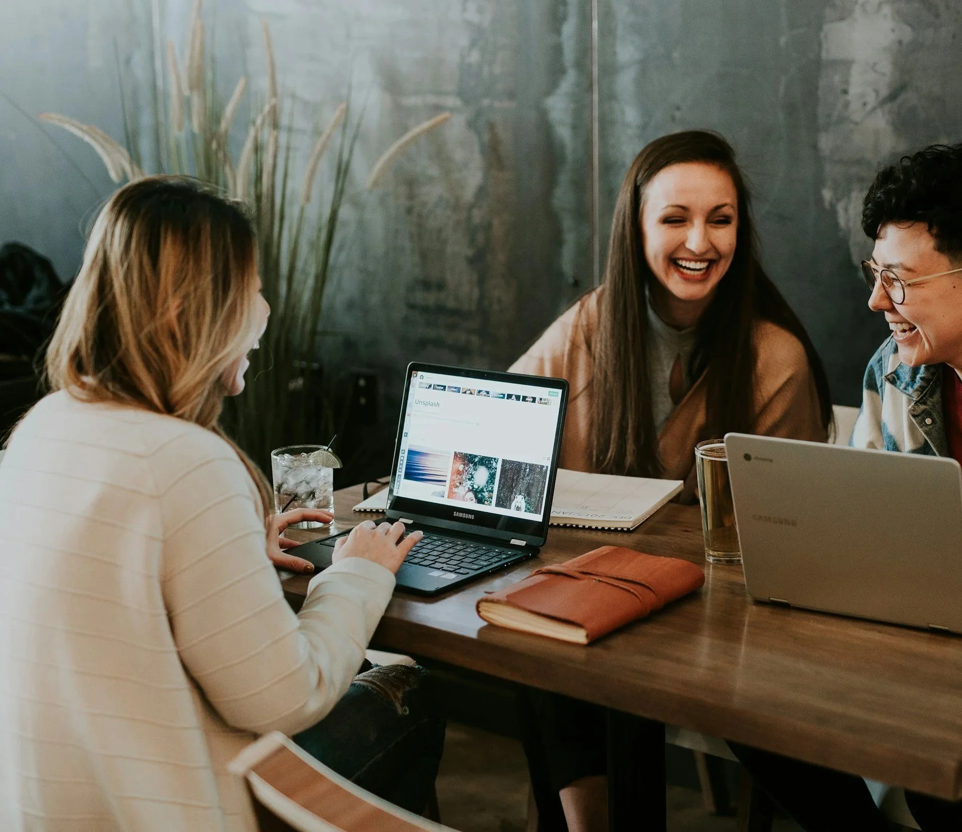 Three people sitting at a table with laptops, smiling and laughing during a meeting in a modern office restaurant.