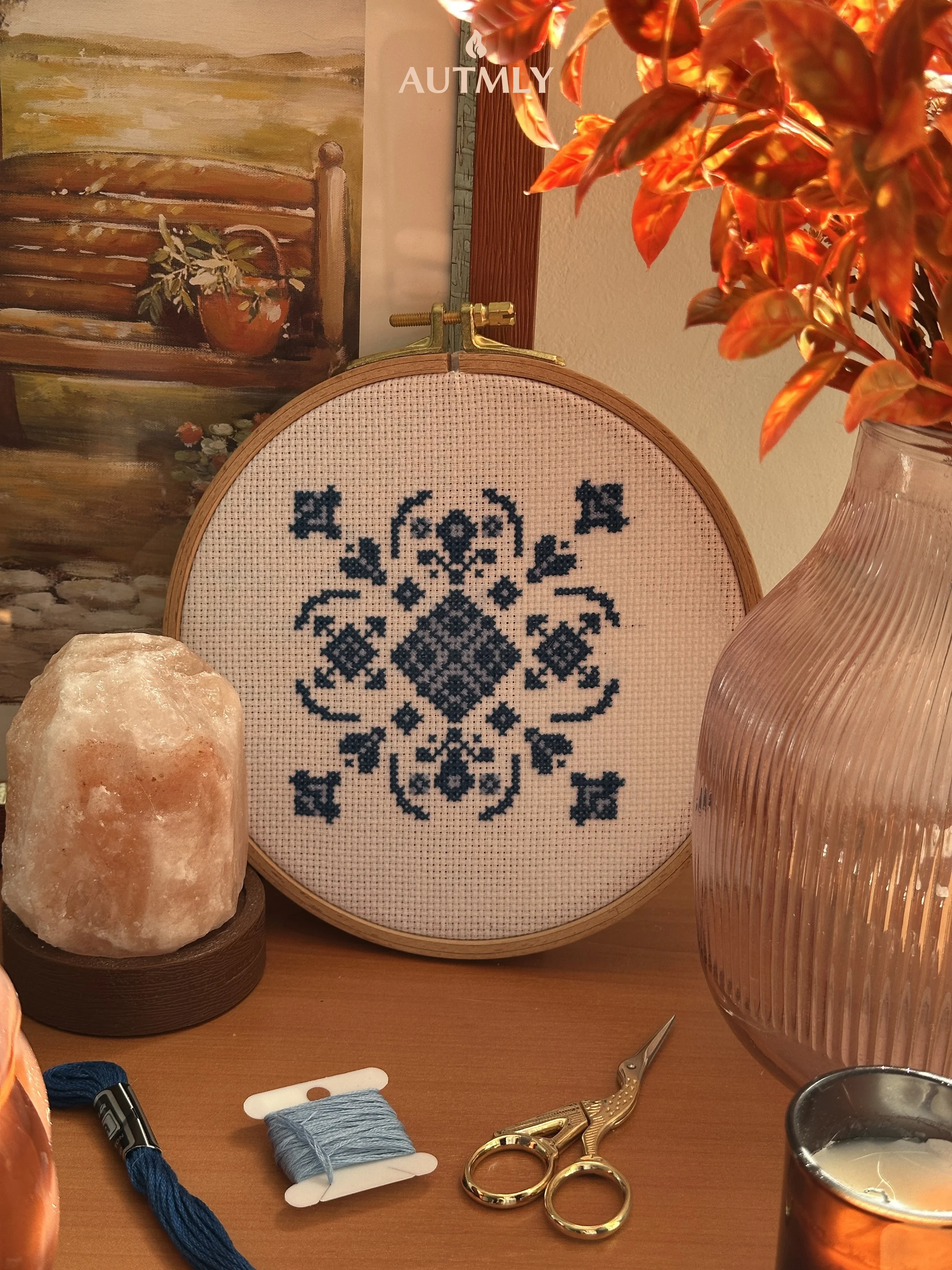 A desk with embroidery supplies, including a cross-stitch hoop with blue fabric and a pattern, a spool of blue thread, gold embroidery scissors, a salt lamp, a pink ribbed glass vase with fake orange leaves, and a candle in an amber glass holder. In the background, there's a framed painting of a pot with greenery and a blanket.