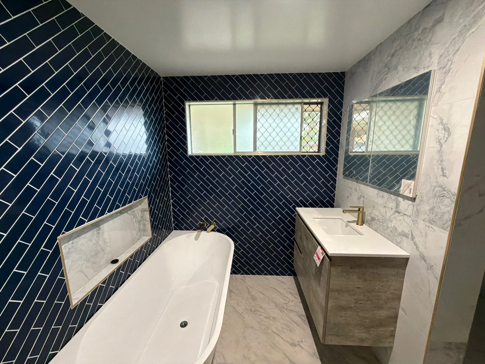 Modern bathroom with navy blue herringbone tile walls, a white bathtub, a vent, a window with frosted glass and security grille, a marble wall with a mirrored cabinet, a vanity with a sink, and gold fixtures.