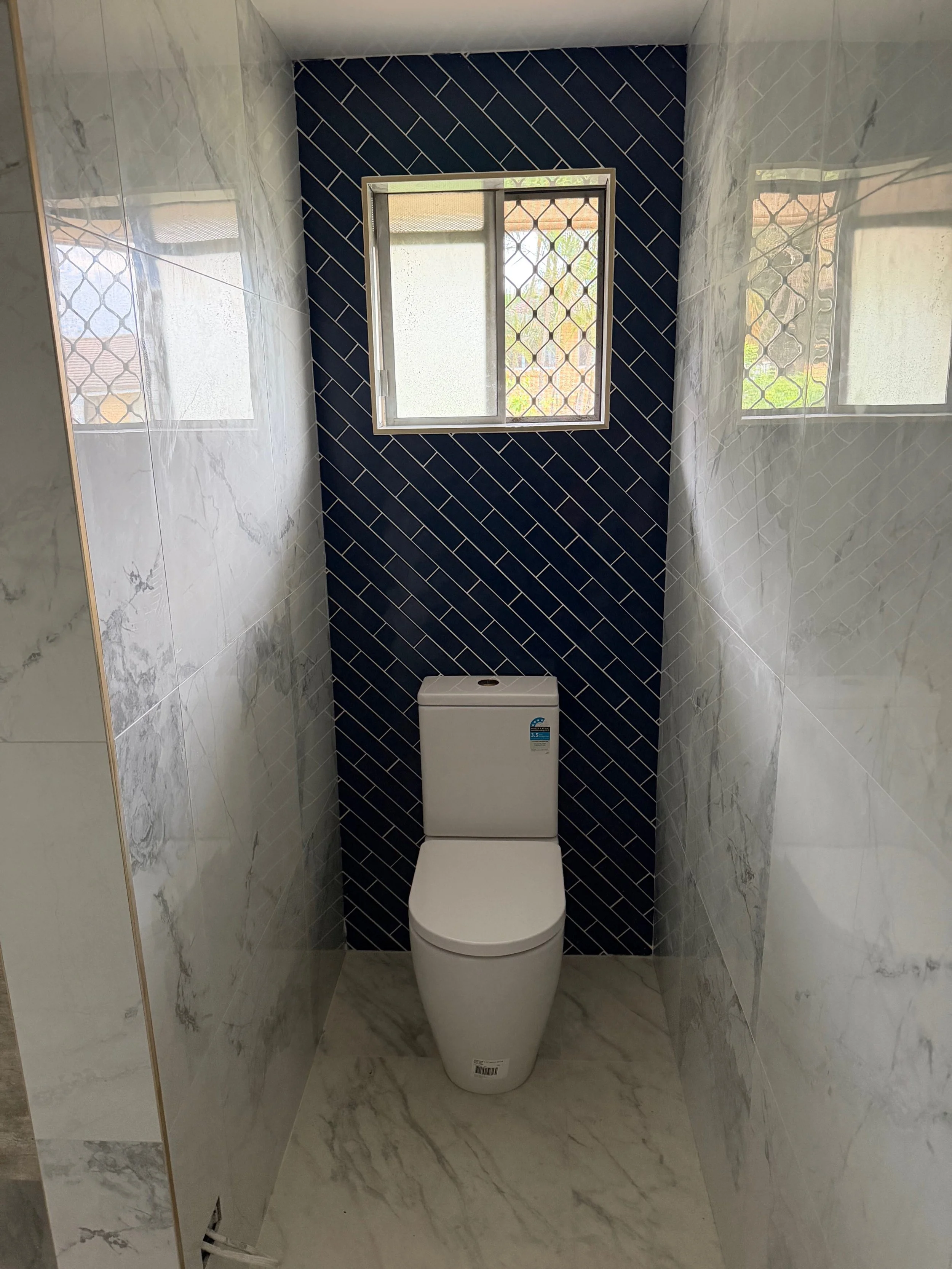 A small modern bathroom with marble walls and floor, a white toilet, a small window with a security grill, and a dark blue tiled wall behind the toilet.