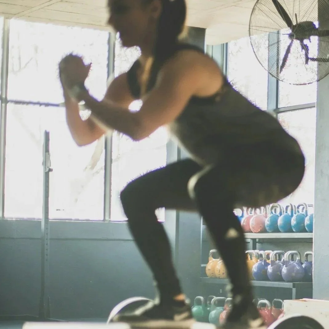 A woman in workout attire performing a squat exercise with a kettlebell in a gym, with colorful kettlebells on shelves in the background and a large fan on the wall.