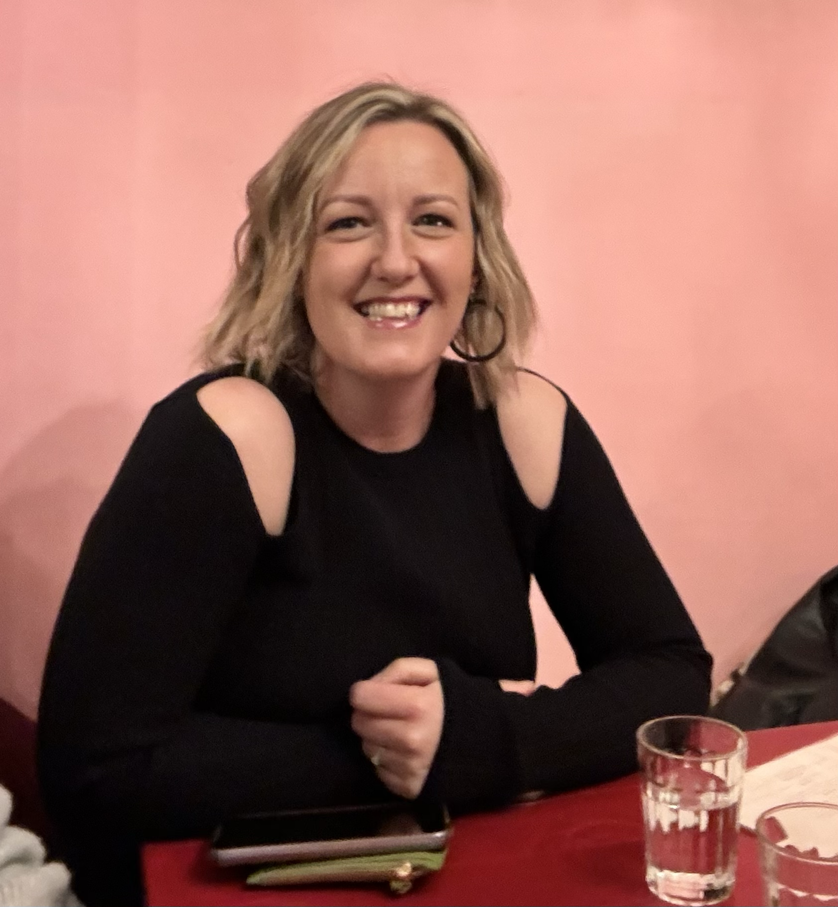 A smiling woman with blonde wavy hair, wearing a black long-sleeve top with shoulder cutouts, sitting at a table with a glass of water and a phone, against a pink background.